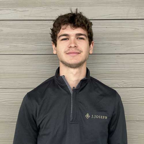 A person wearing a dark pullover with "J. Joseph" embroidered on it, standing in front of a wooden wall.