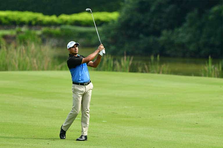 A golfer mid-swing on a green golf course, wearing a blue and black shirt, beige pants, and a white cap. Trees and bushes are visible in the background. This is how to hit a 50 yard pitch shot.